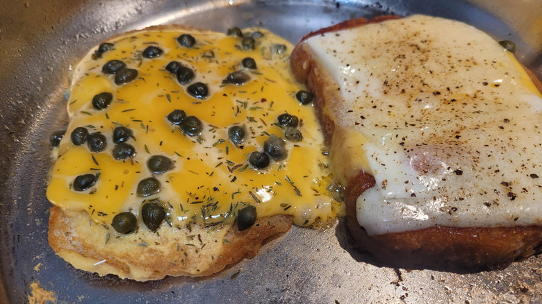 two slices of bread cooking in a skillet, both topped with melted cheese, and one with capers as well