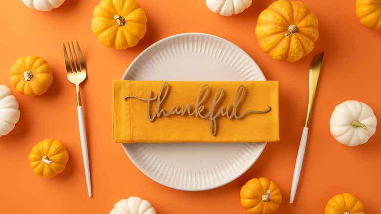 Thanksgiving table setting with small pumpkins, paper plate, and orange napkin that reads "thankful"