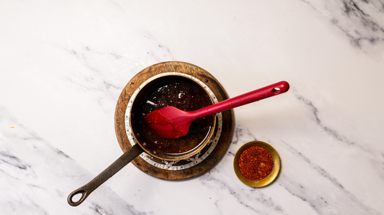 Hot honey in saucepan with spatula besides pinch bowl with ground Thai chile flakes.