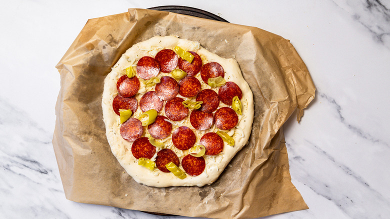 Dough with cheese, pepperoni and pepperoncini spread out on parchment paper.