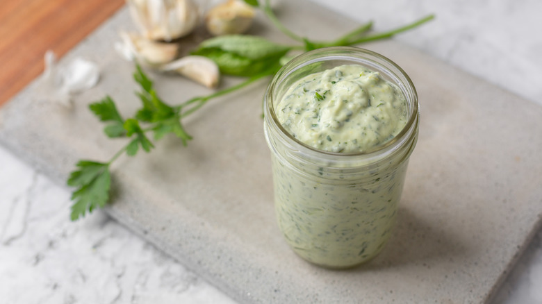 Jar of creamy basil dressing next to sprigs of parsley and garlic cloves