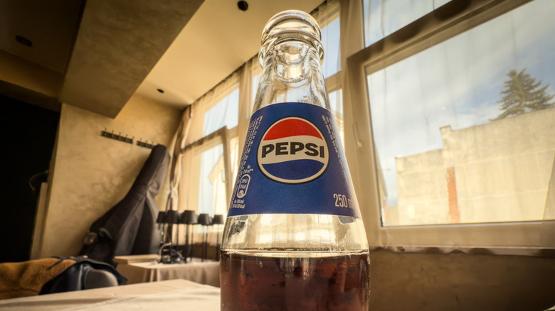 A partially full glass bottle of Pepsi in front of a window