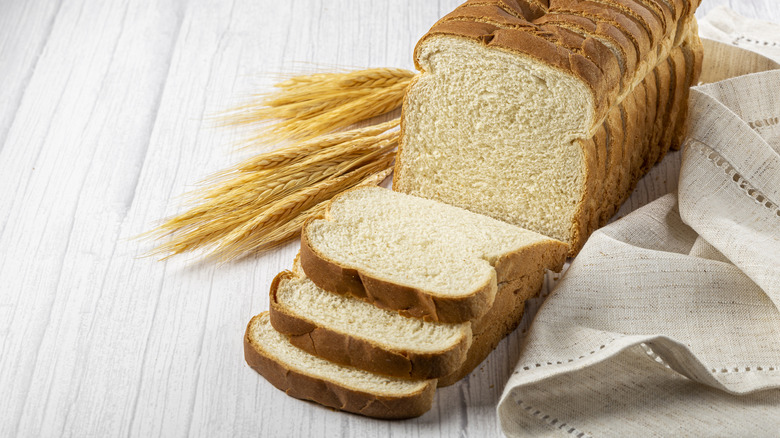 Sliced loaf of white bread on white surface next to wheat grains and white cloth