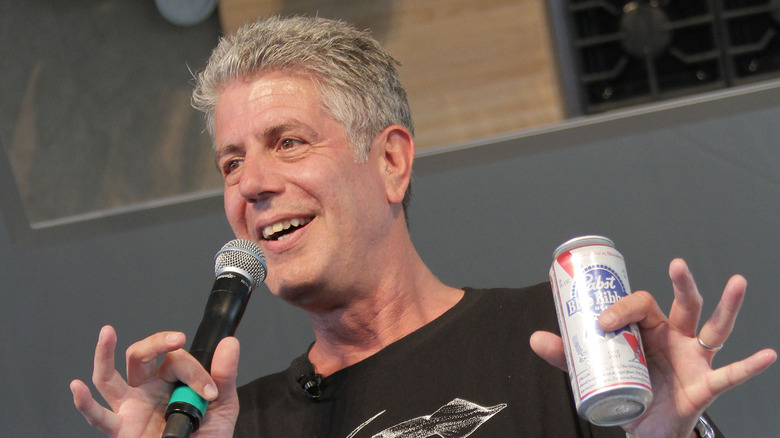 Anthony Bourdain in a black T-shirt holding a microphone in one hand and a Pabst Blue Ribbon beer in the other