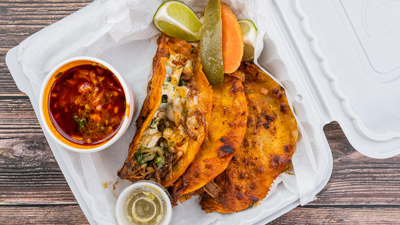quesabirria tacos in a to-go container with a cup of consomé on the side