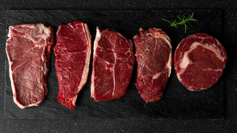 5 different cuts of steak on a slate with a sprig of rosemary