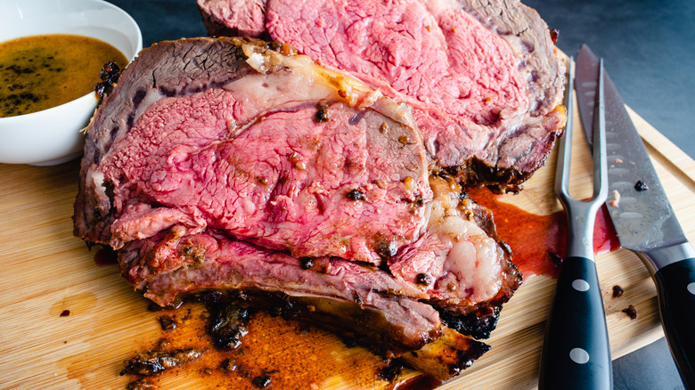 Two carved pieces of prime rib on a wooden cutting board with prominent fat caps (layers of fat along the top of the meat). They're served alongside a bowl of jus.