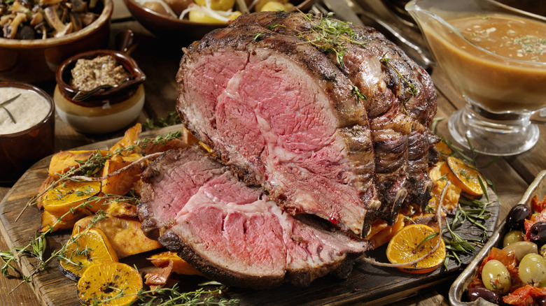 A medium rare prime rib on a cutting board with several citrus slices, surrounded by fixings like gravy and a platter of olives.