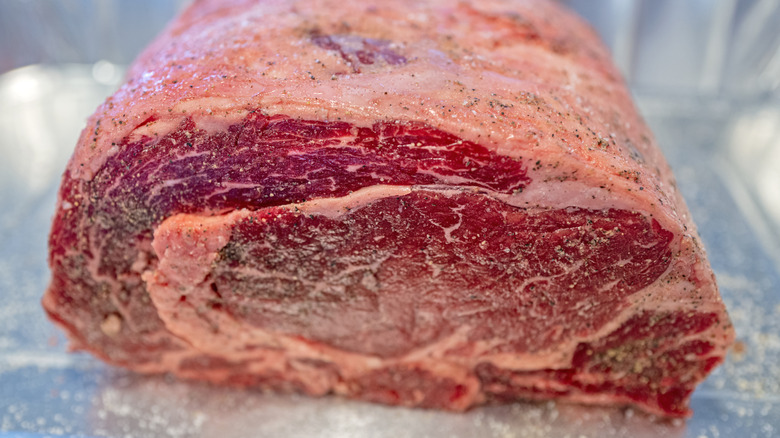 A bright red piece of raw prime rib, seasoned with salt and pepper, sitting inside an aluminum roasting pan.