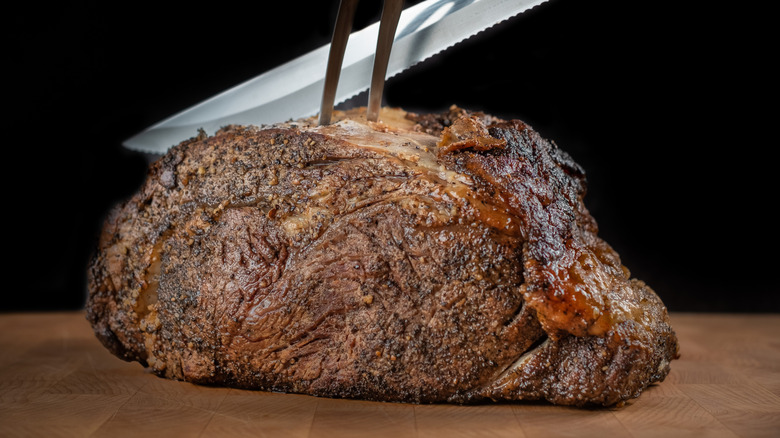 A serrated knife cuts into a browned piece of prime rib on a wooden cutting board.