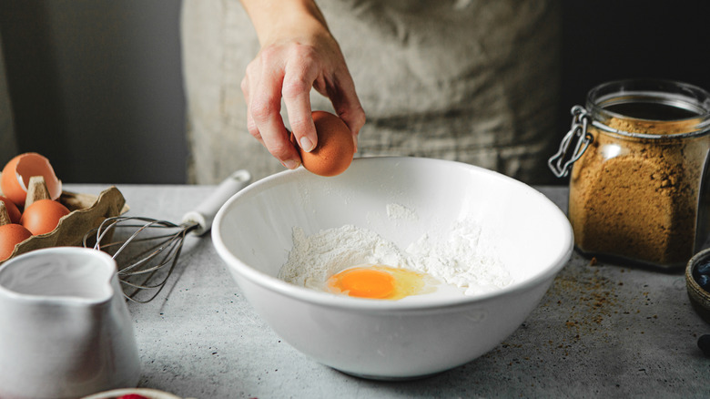 A hand cracking one egg on the side of a white bowl