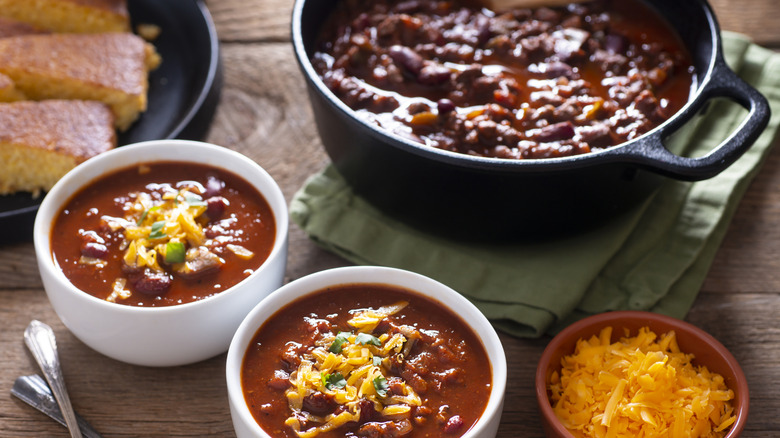 Two bowls of chili rest on a wooden table next to a large crockpot of chili, a smaller bowl of cheese, and a plate of cornbread.