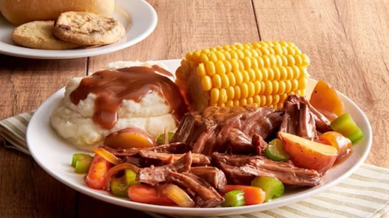 A plate of gravy covered pot roast with potatoes, celery, and carrots sitting on a plate with gravy-covered mashed potatoes and corn on the cob
