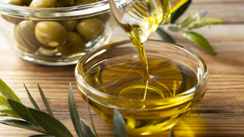 Olive oil being poured into a glass bowl with a glass bowl of olives in the background.