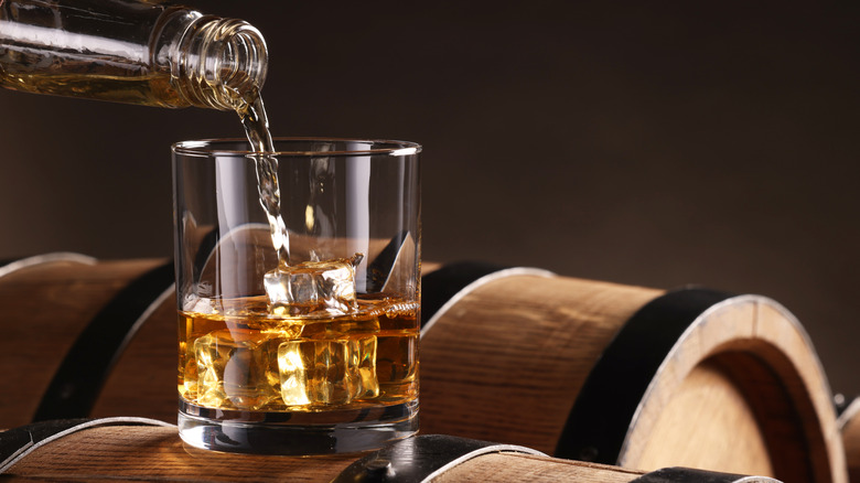 Whiskey being poured from a bottle into a glass set atop an oak barrel