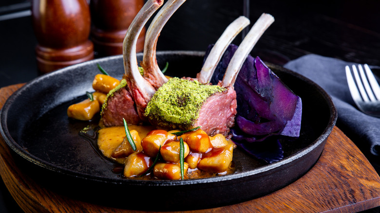Double-cut lamb chops on plate with colorful sides on a black plate with wooden salt and pepper mills in the background and a fork and napkin nearby.