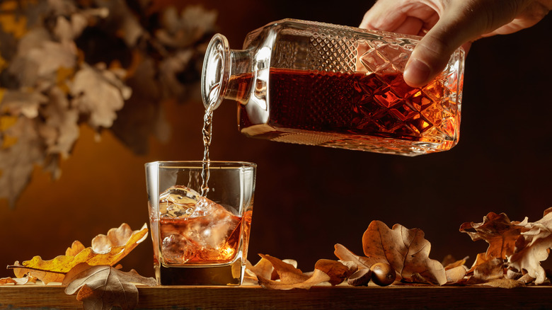 Brown liquor poured from a crystal bottle into a glass of ice next to dried leaves