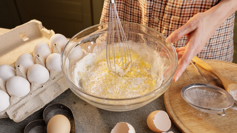 a person mixes cake batter in a glass bowl, with eggs and other tools scattered around the table