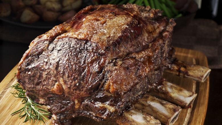 A fully cooked prime rib roast rests on beef bones atop a wooden carving board, with side dishes of potatoes and green beans in the background