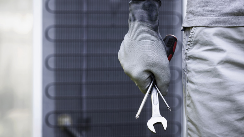 A repair technician standing in front of the back of a fridge holding a spanner and screwdrivers in a gloved hand