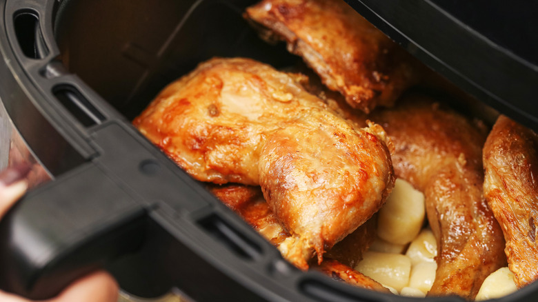A person opening the  basket of an air fryer to check the chicken and potatoes cooking inside