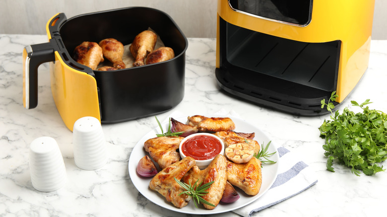 A small yellow air fryer with its basket full of chicken drumsticks, on a counter next to a white plate of chicken and sauce