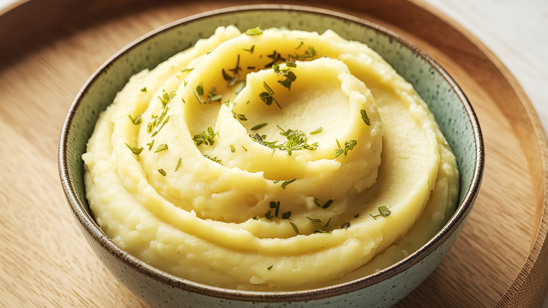 Mashed potatoes in a bowl with herbs next to garlic, herbs, spuds, and cream