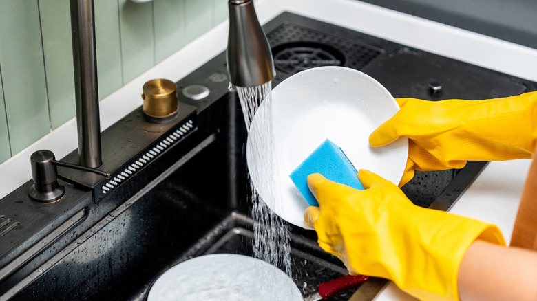 Gloved hands washing dish under running faucet in sink