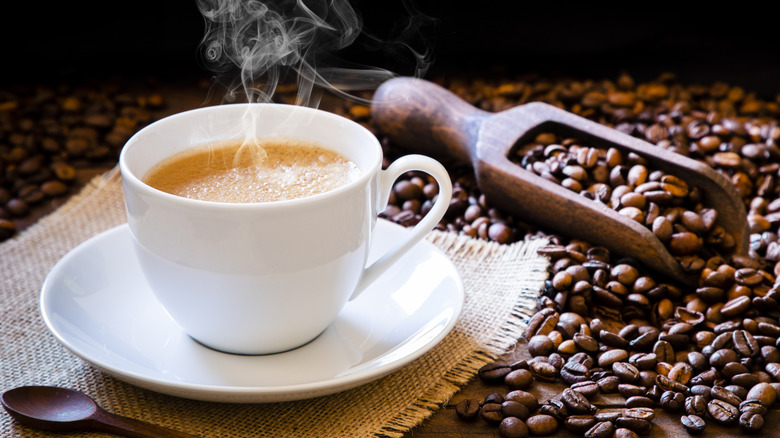Steaming white mug of coffee on saucer next to coffee beans and wooden scooper in dark room
