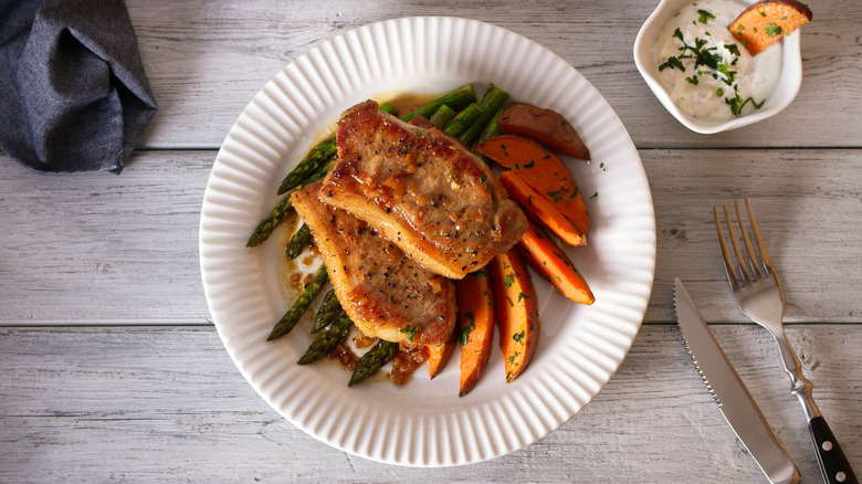 Pork chops with sweet potatoes on plate next to cutlery and bowl of white sauce
