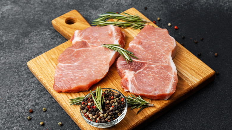 Raw pork chops on wooden board next to rosemary sprigs and peppercorns