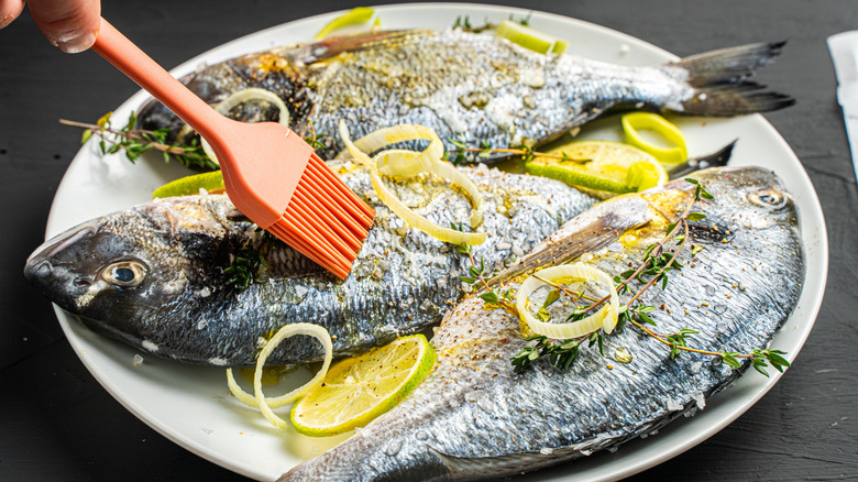 Raw whole fish with lime slices, thyme, and someone brushing on oil