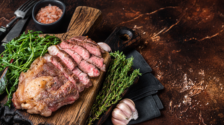 A chuck roast sits on a cutting board surrounded by fresh herbs, shallots and salt. There is a black napkin and cutlery nearby