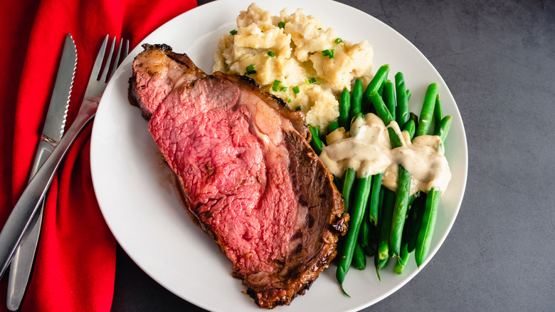 A cut of rare prime rib next to bright green beans ith gravy and mashed potatoes with chives.