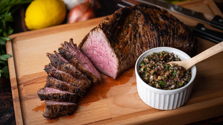 A grilled tri-tip steak on a wooden cutting board with a topping beside it in a porcelain ramekin. There are shallots and lemons back and to the side