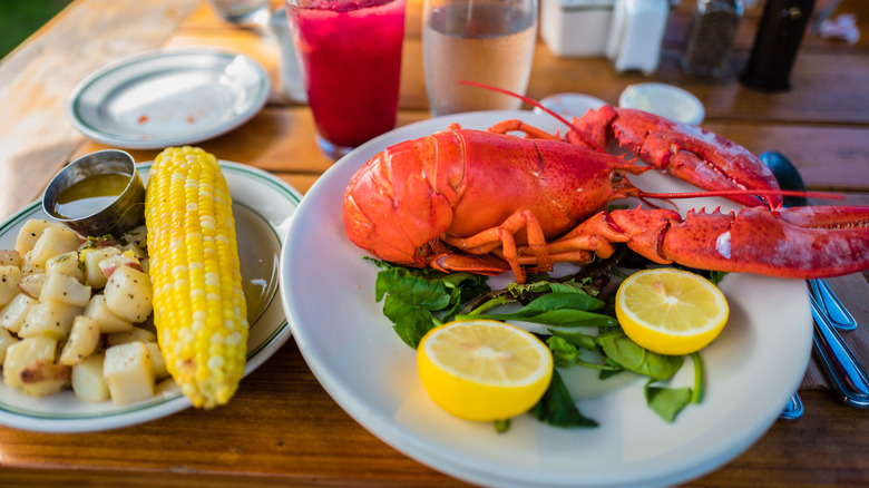 Whole cooked lobster on a plate with greens and lemon next to a plate with corn and potatoes with drinks in the background.
