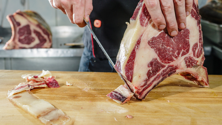 Someone trimming the fat off a large, dry aged ribeye on a cutting board