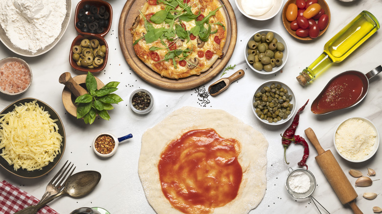 Pizza making supplies, finished pizza, dough with sauce, olives, ,tomatoes, cheese, and capers in bowls, olive oil and various utensils