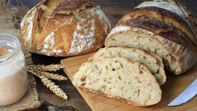 a whole loaf and a sliced loaf of sourdough bread next to a sourdough starter in a glass jar