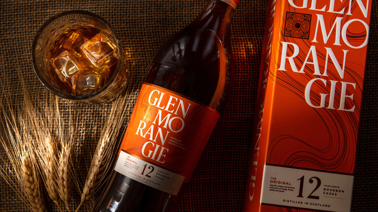 A glass of Glenmorangie on ice beside barley, the bottle, and its box