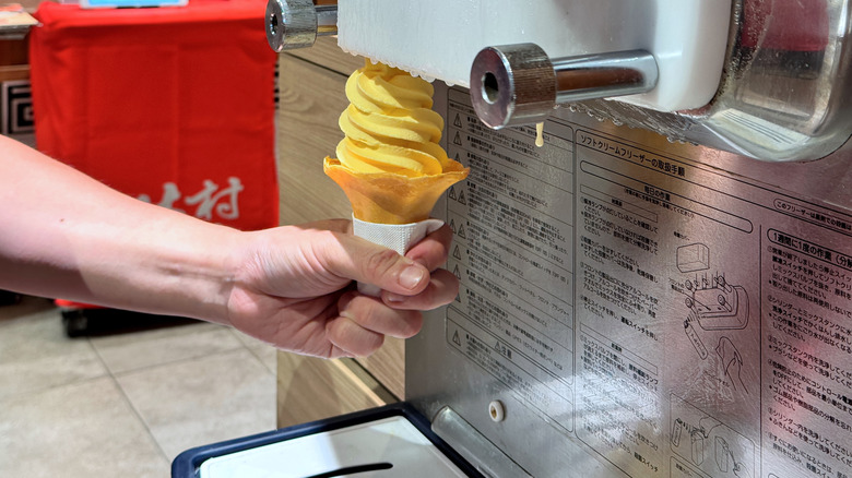 Yellow soft-serve ice cream cone held beneath the nozzle of a soft-serve machine.