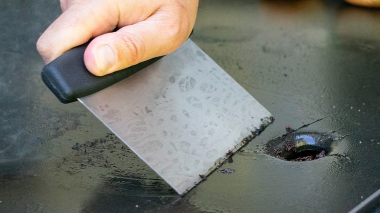 Hand holding metal scraper and scraping it across black surface of griddle