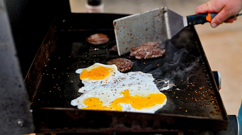 Person cooking fried eggs and hamburger patties on Blackstone griddle using flat metal spatula