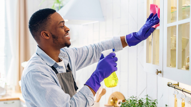 Man wearing purple dish gloves wiping white cabinets with pink cloth