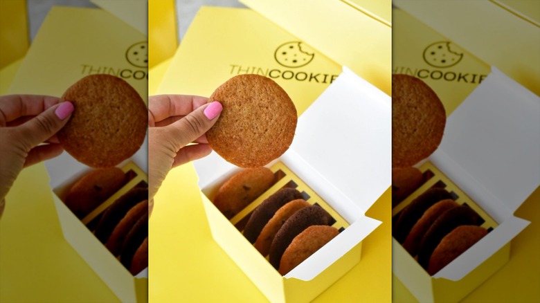 A yellow box of Thin Cookies with someone holding up a single cookie