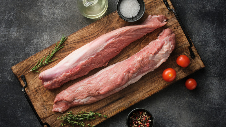 two raw pork tenderloins on a wooden cutting board surrounded by herbs and spices