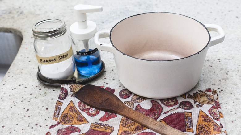 White Dutch oven with baking soda, dish soap, cloth and wooden spoon