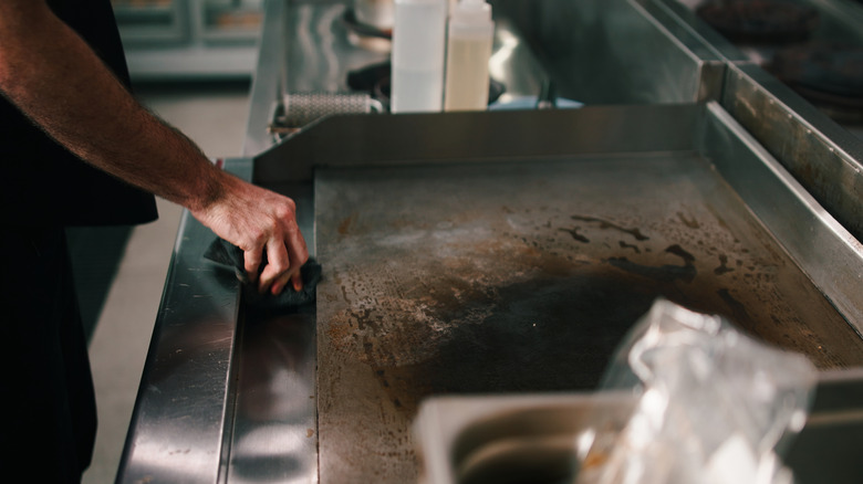A man standing over a flat-top grill cleaning it with a large scouring rag in a professional kitchen