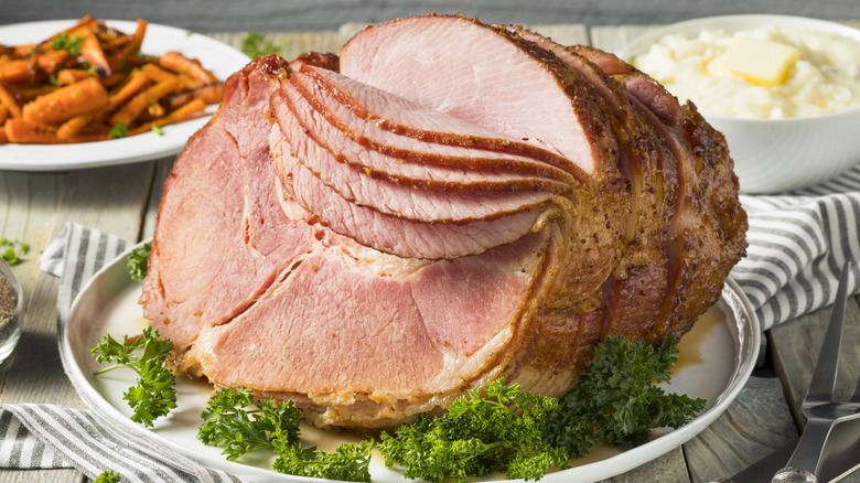Roasted and partially sliced ham on a plate with some parsley garnish and side dishes in the background