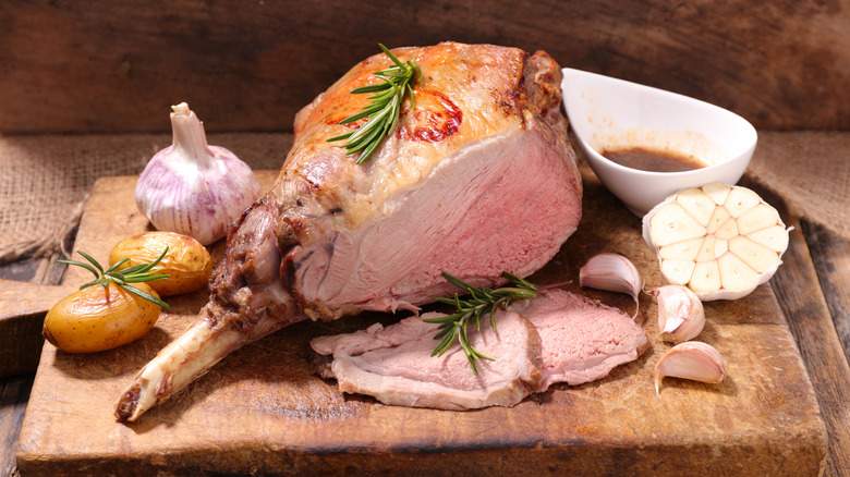 a bone-in roast leg of lamb sitting on a cutting board with rosemary, potatoes and full bulbs of garlic